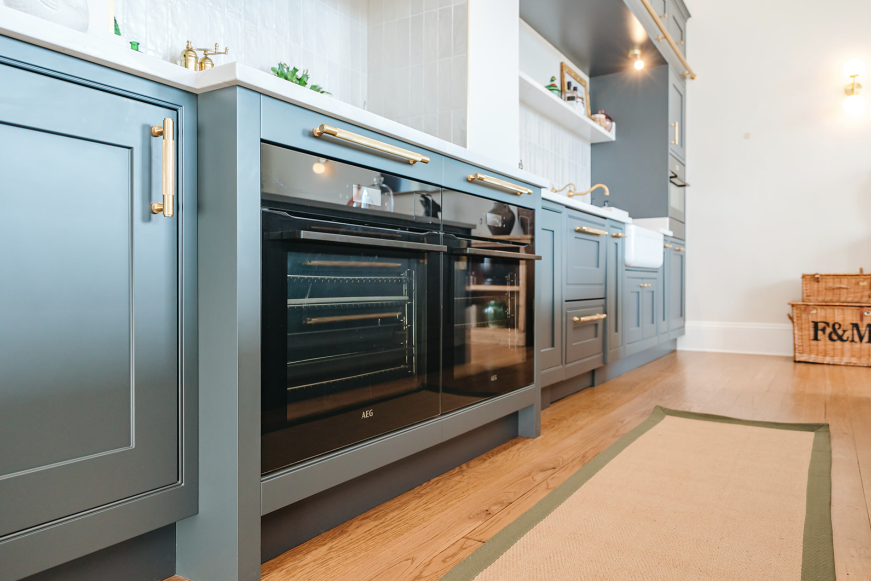 A run of blue-grey inframe cabinetry with twin ovens, brass bar handles and a butler sink beyond