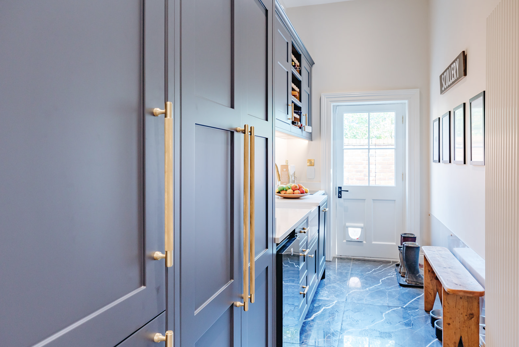 The Bicton Hall scullery: blue-grey inframe cabinetry with brass handles, marble floor, boot bench and a half-glazed door to the garden