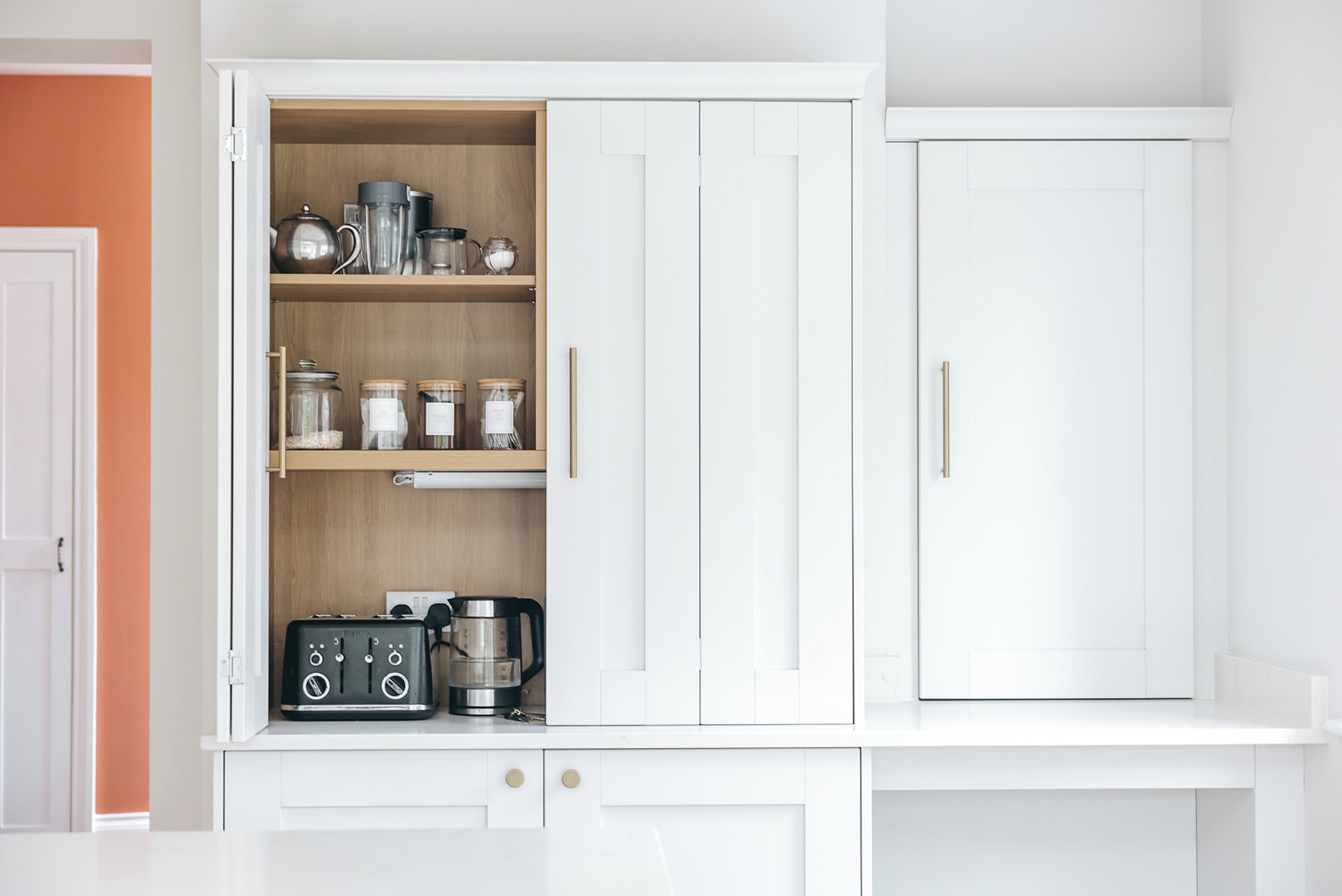 White bi-fold appliance cupboard opened to reveal an oak-lined interior holding a toaster, kettle and storage jars