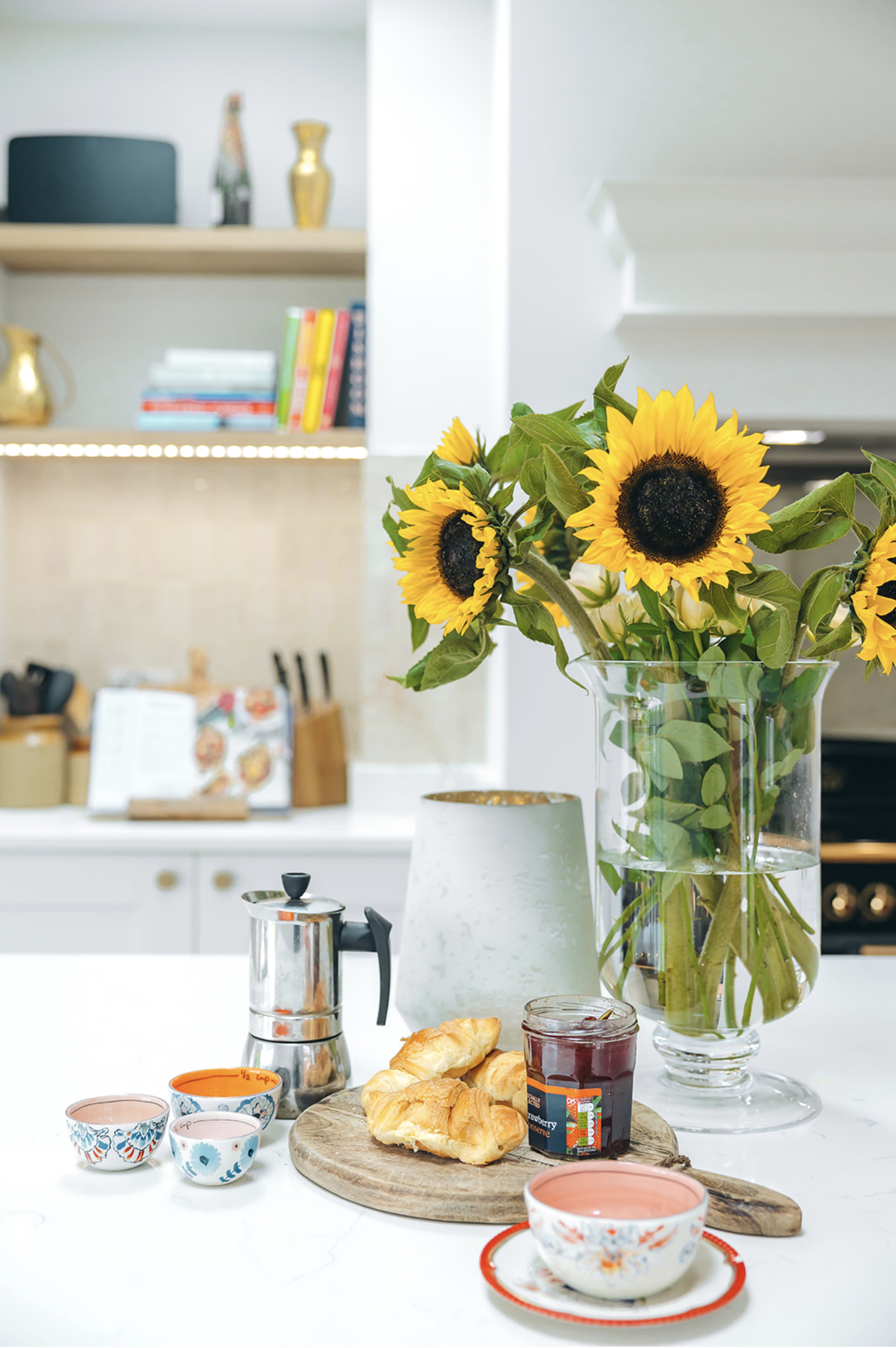 Kitchen island styled with sunflowers, a stovetop coffee pot and open shelving with cookbooks behind