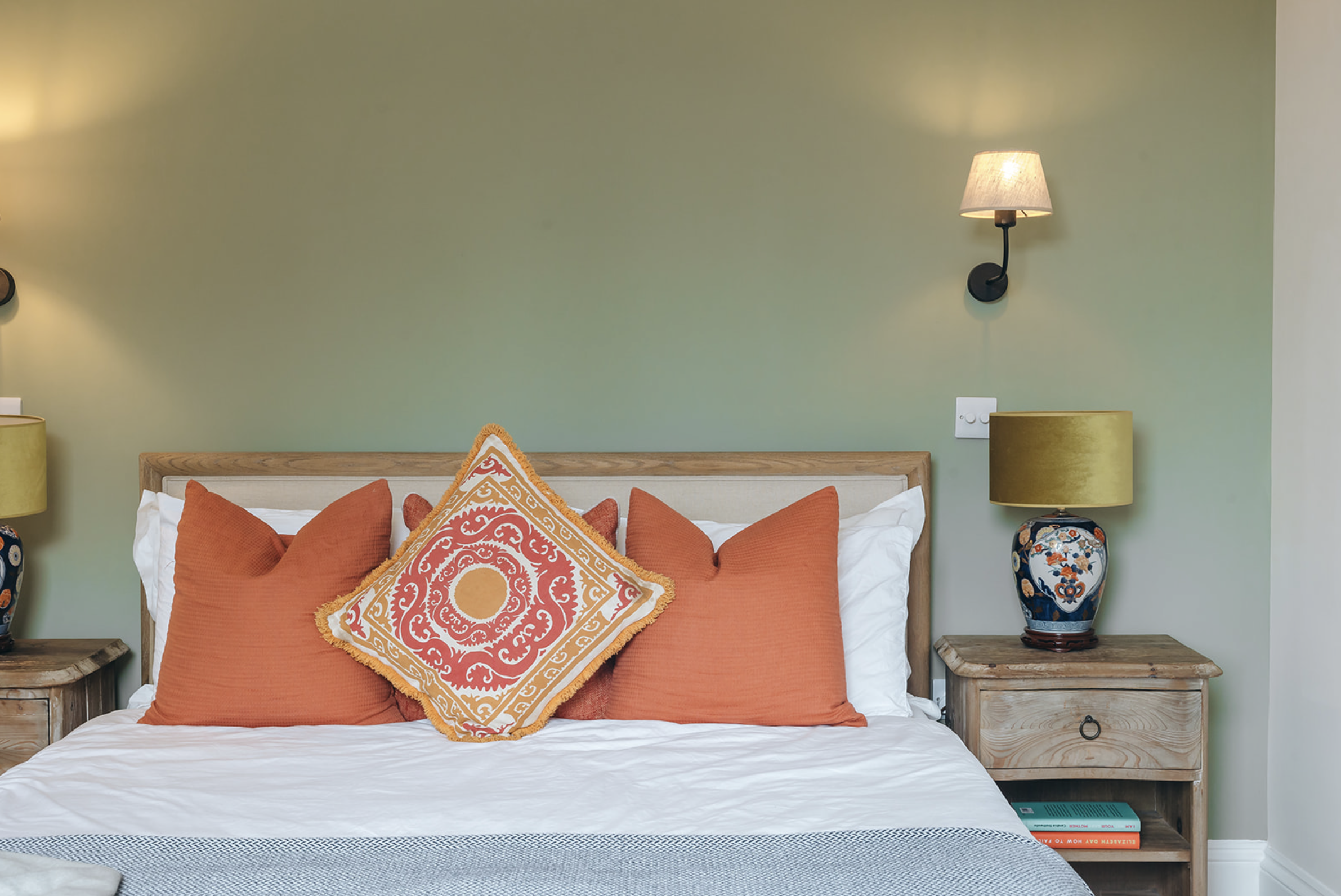 Bedroom with an oak-framed upholstered headboard, orange cushions and brass wall lights against a soft green wall