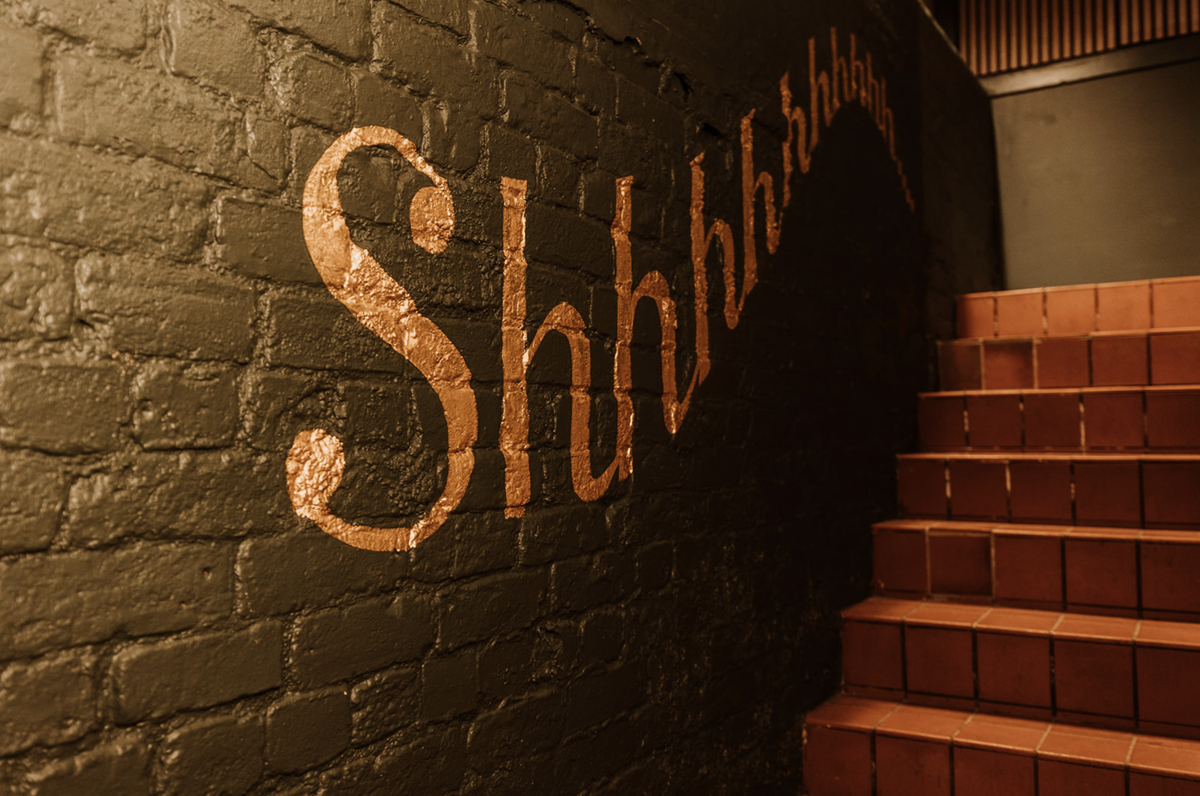 Gold-leaf Shhh signage hand-painted on the dark brick wall of the stairwell