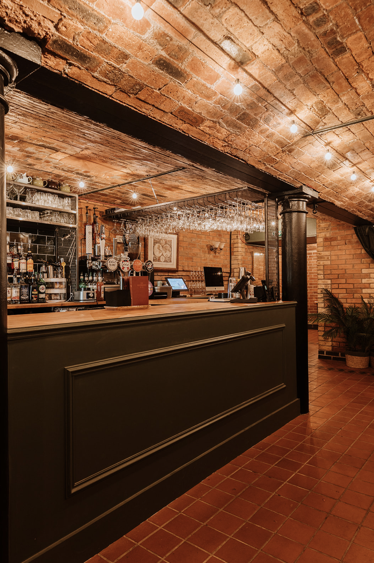 The Underground bar: green panelled counter, timber top, hanging glass rack and cast-iron column beneath the brick vault
