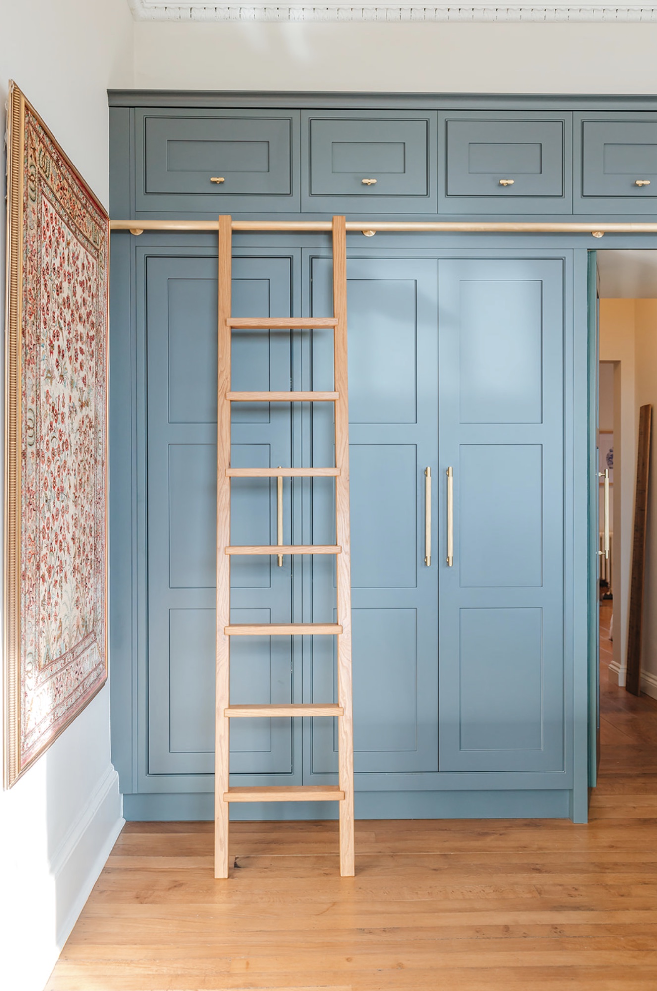 Oak library ladder against full-height teal kitchen cabinetry with brass rail and handles