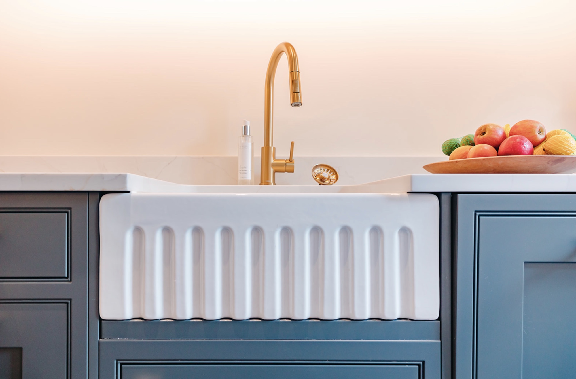 White fluted scullery sink with a brass tap, dark teal cabinetry and a bowl of fruit