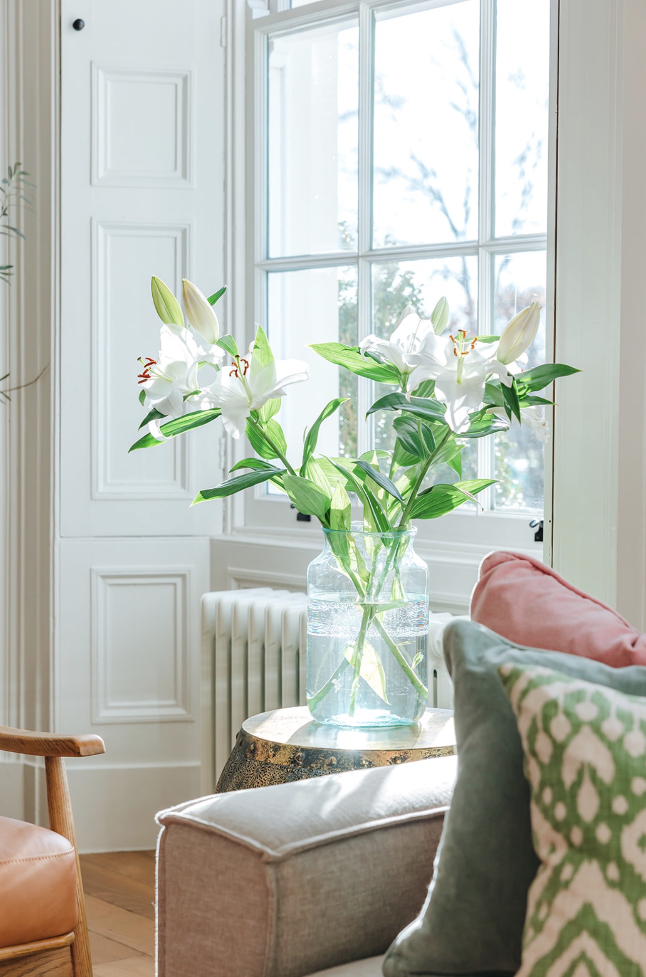 White lilies on a side table beside a shuttered sash window and layered green cushions