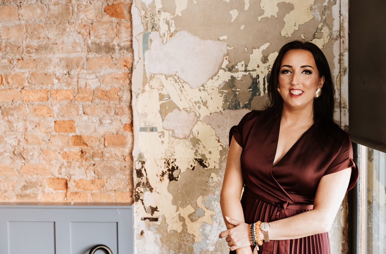 Camilla Monk smiling, standing against a stripped brick and plaster wall