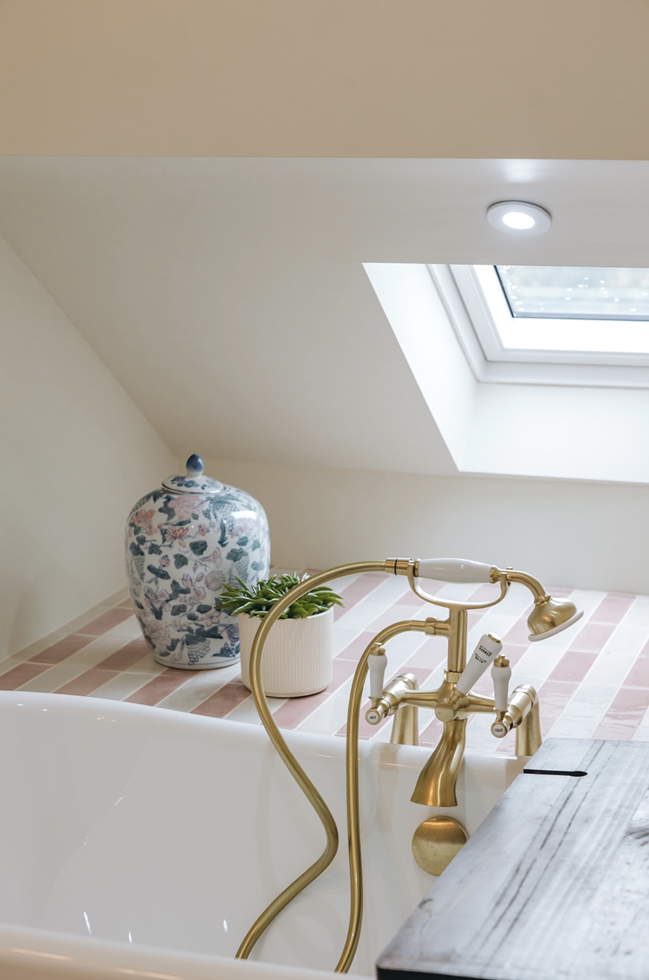 Freestanding bath with brass telephone tap beneath a rooflight and pink striped tiled ledge