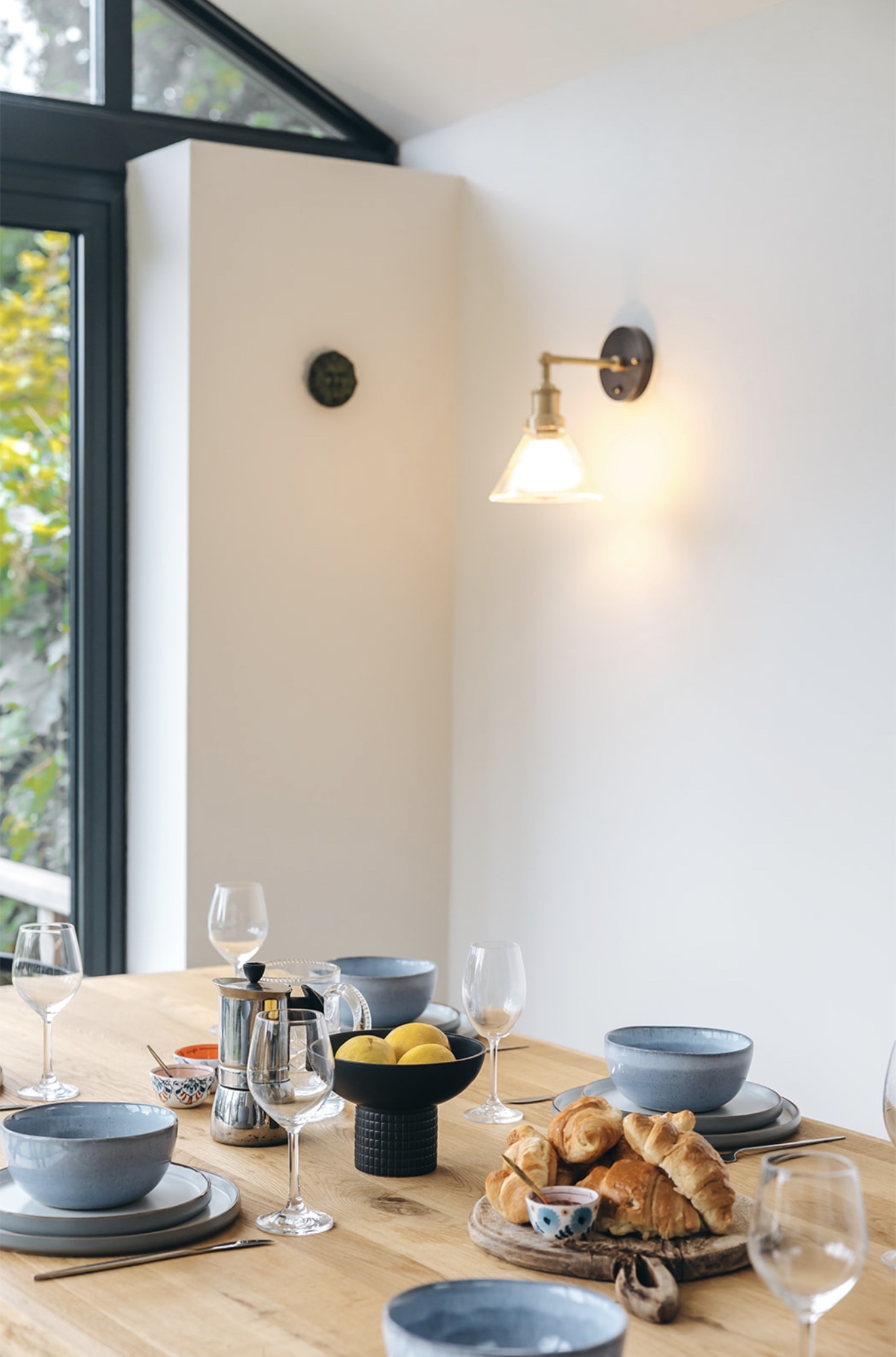 Oak dining table set with blue ceramics, glassware and breakfast beneath a brass wall light