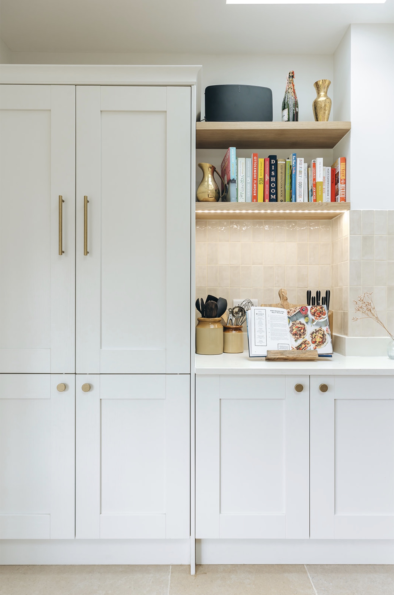 White shaker cabinetry beside an oak-shelved pantry niche with cookbooks, glazed tiles and concealed lighting