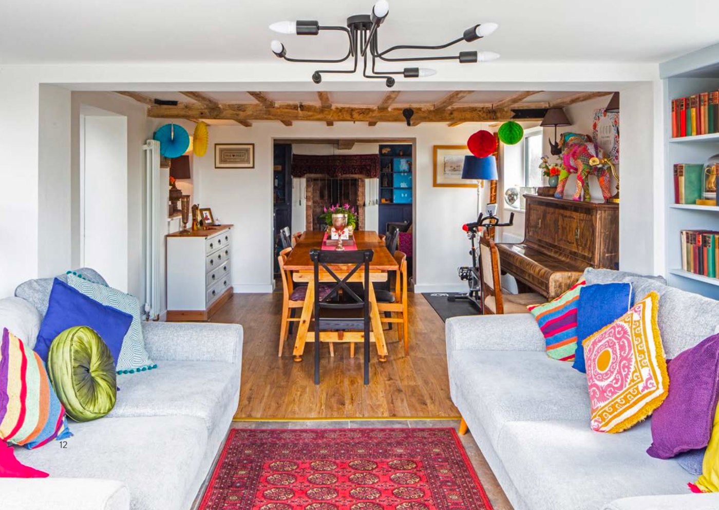 Mount Pleasant open-plan sitting and dining space beneath exposed oak beams, with grey sofas, a red rug, piano and a view through to the inglenook fireplace beyond