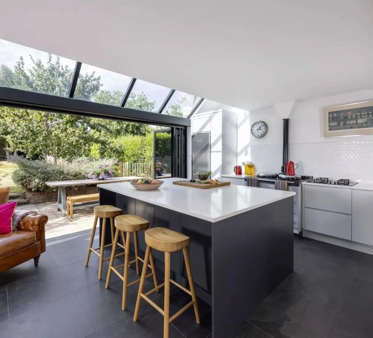 Oakleigh House kitchen: dark island with oak stools beneath a fully glazed roof, white units and bifold doors open to the garden