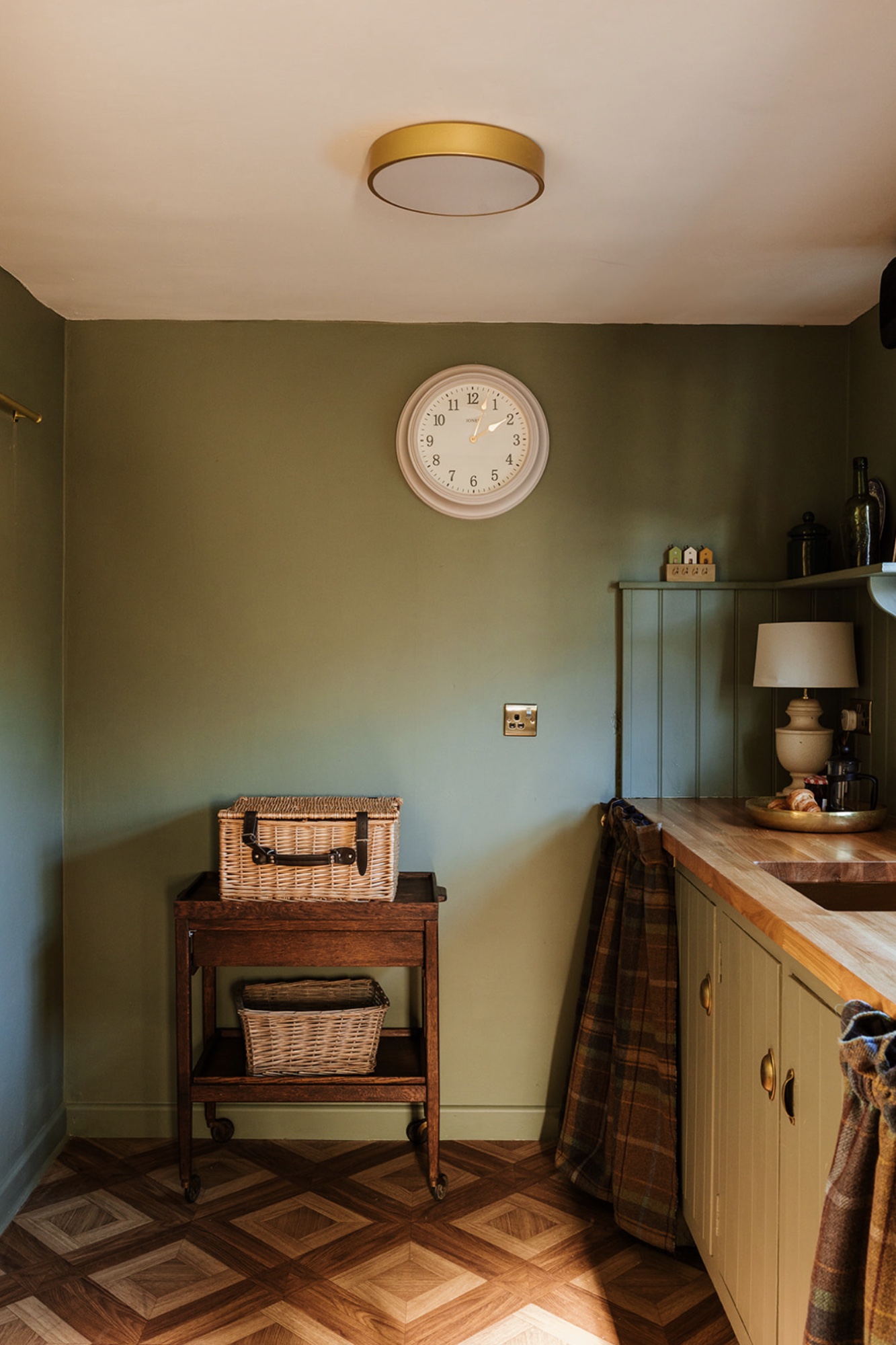 Pool House kitchenette corner with a wall clock, wicker baskets and green cabinetry